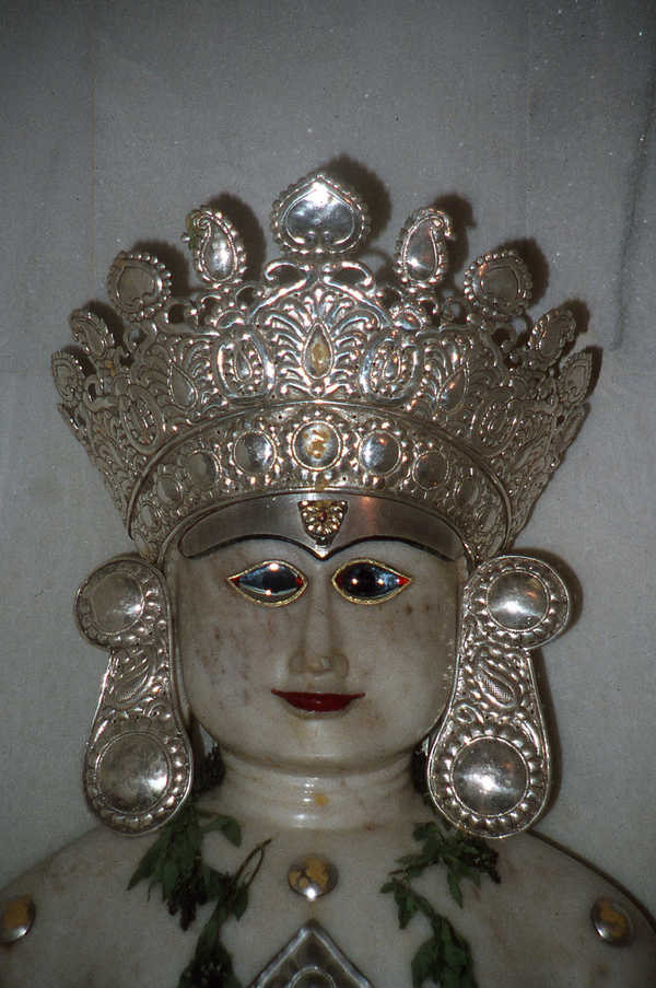 The head of white stone sculpture is crowned with a large, patterned metal crown and oversized earrings.The face has shining glass blue eyes that have black irises and red tips. 