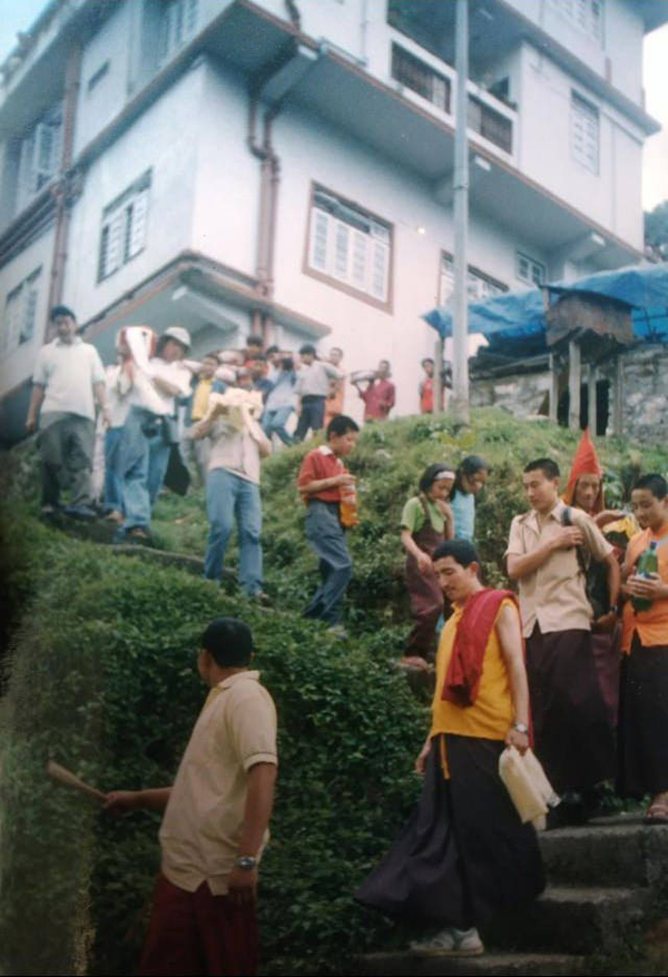 A line of South Asian men and women process down a winding set of steps. Some carry scarf-wrapped books on their shoulders.