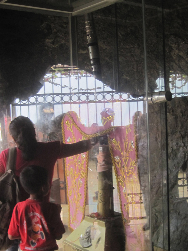 A glass sheet protects a wooden cross that has been decorated with a purple stole. A woman reaches through a hole in the glass to touch the cross while a young boy looks on.