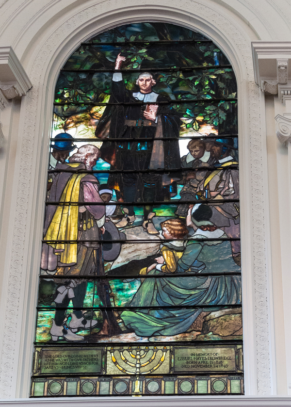 A luminous stained glass window depicts a group of figures in Puritan garb gathered around a minister. He stands with his arm raised in oration.