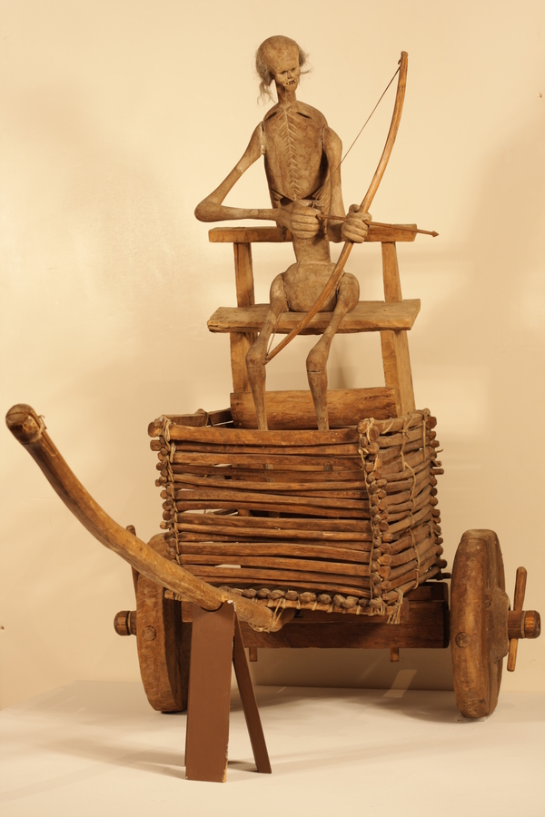 A square wooden cart on two wheels holds a chair with a large skeletal figure. The cart stands on two wheels with a large, protruding chassis. The skeletal figure looks out sideways from the cart and pulls a bow and arrow taunt. 