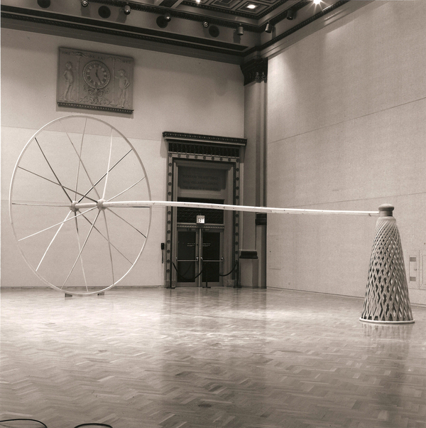 A black-and-white photo shows a massive wooden sculpture installed in a neoclassic interior. A pole joins an oversized wooden wheel to an large, oblong inverted woven basket. 