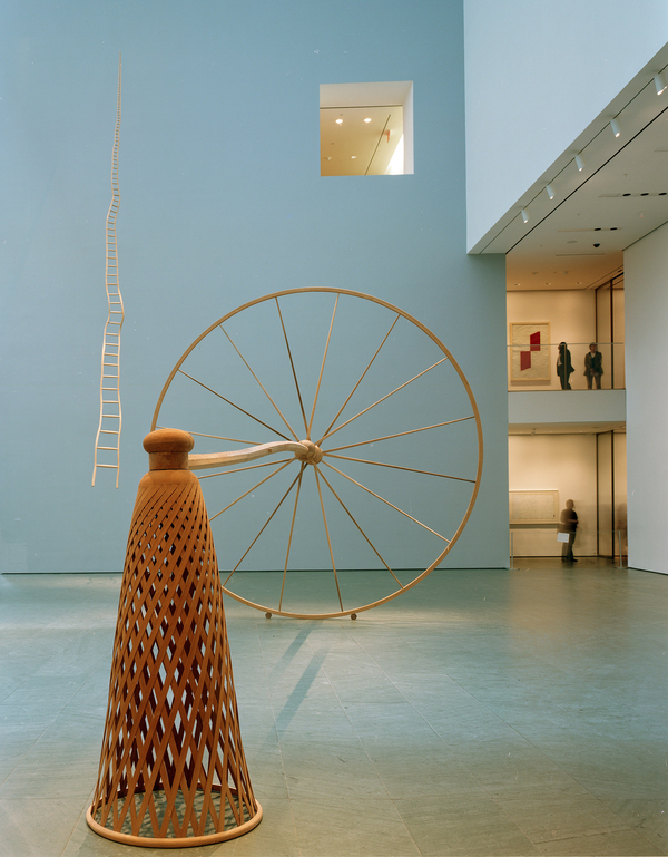A tall, inverted wooden basket connects to a massive wooden wheel via a long, horizontal wooden pole.