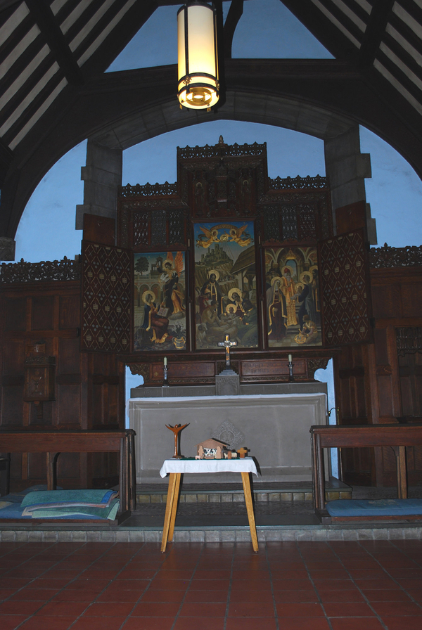 A tripartite altar screen opens to a panel painting of the Annunication, Nativity, and the Presentation at the Temple. It is part of a larger wooden reredos behind a stone altar. A small table with a nativity set stands in front of the altar and screen.