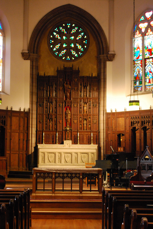 A photograph down a church aisle shows a white altar with a large wooden altar screen. Wooden icons are embedded in the screen. It stands under a stained glass rose window. 