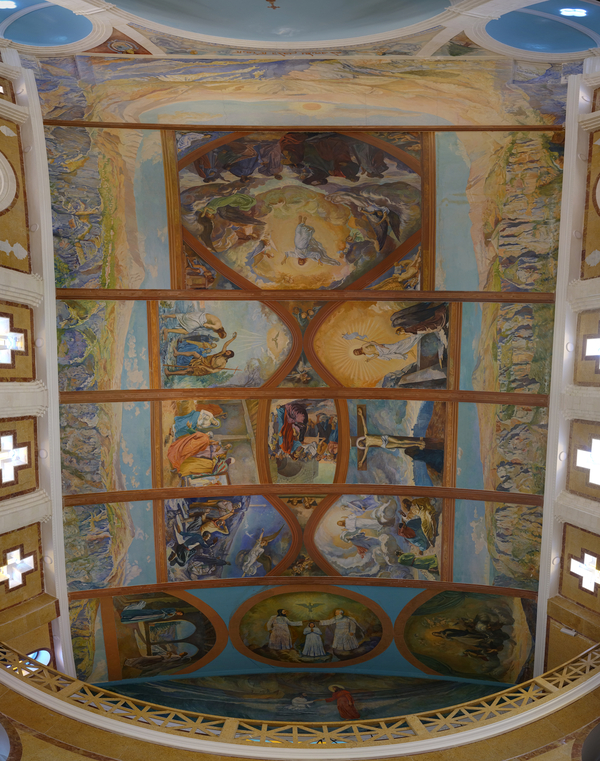 The mural paintings on a church ceiling depict brightly colored biblical vignettes down a central vertical axis. Mountain ranges are painted on the other sides. Wooden ribbing separates the scenes.