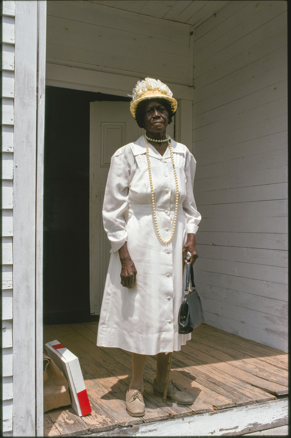 An older black woman stands on a wooden porch in a color photograph. She wears a buttoned white dress, a decorative white hat, and large pearls. She holds a black handbag and looks out at the camera seriously. 