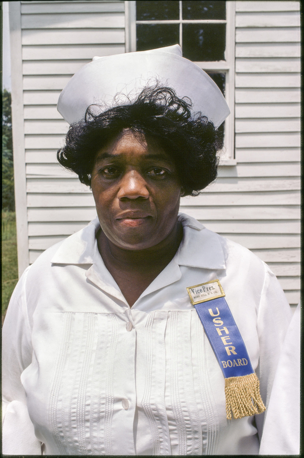 A tight portrait of a middle-age black woman captures her white dress and crisp white hat on curled hair. She wears a blue badge that reads "Vice Pres. USHER BOARD." 