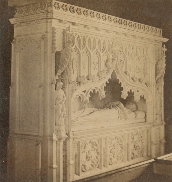 This black and white photo captures a marble canopy tomb from a diagonal. The relatively undecorated side of the monument, which contrasts with the heavily carved canopy and tomb chest, protrudes from the wall. 
