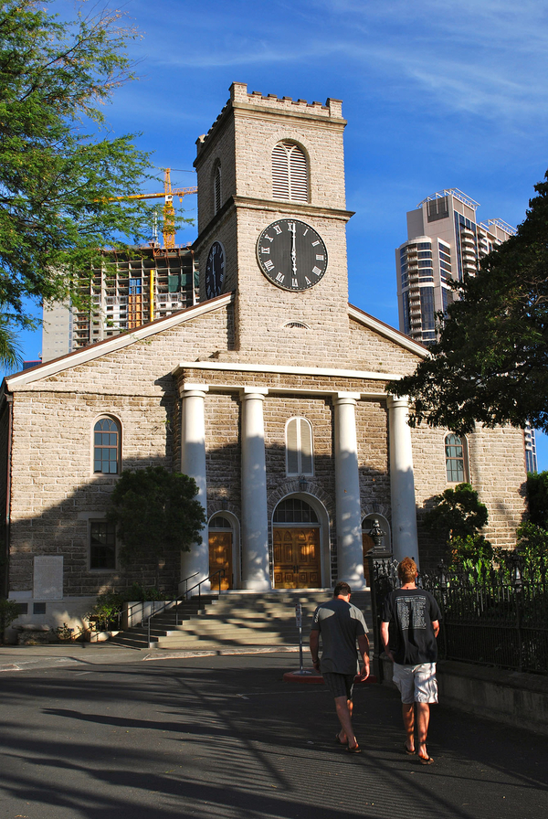 People walk towards a light-colored, coral brick church. It has a pedimented facade with a collonated entryway and a large, crenellated tower on top. Modern buildings are visible in the background.