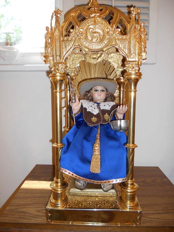A light-skinned figure of a young boy wears a shiny blue robe, brown stole, and wide-brimmed hat. He is seated in a gold throne with an ornate canopy carved with a lamb insignia. The child holds a basket and staff.