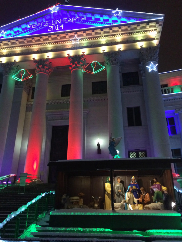 A nativity scene sits in front of a white colonnaded civic building that is lit with neon lights. They are shaped like bells, stars, and a sign that reads "Peace On Earth 2014." 