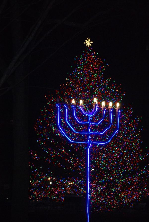 A blue neon menorah with six lit lights stands in front of a multi-colored lit Christmas tree.