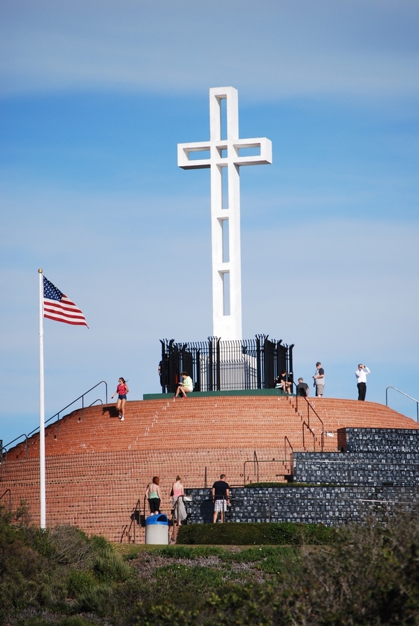 People climb up a tall stepped platform that bears a very large white concrete cross. The cross is constructed out of blocks that outline the cruciform shape but leave empty space in the center.