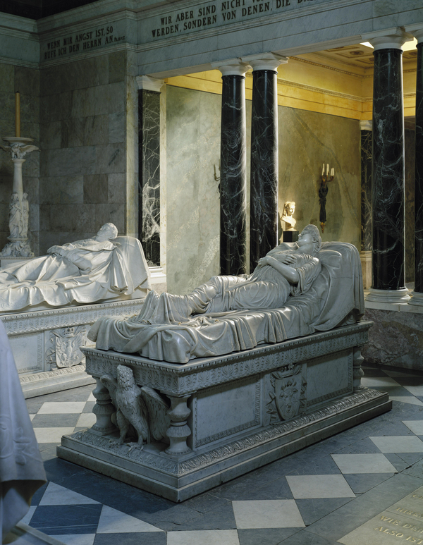 Two marble sarcophagi are visible in a marble mausoleum, both with a recumbent effigies on theirs lids. The female figure reclines with her hands and legs crossed naturally, as if asleep on a plush cushion. Flowing robes cling to her body. 