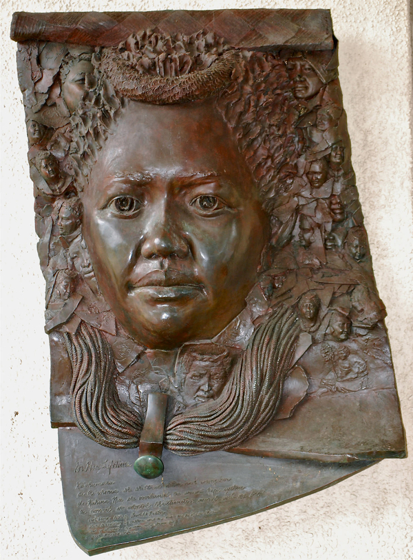 A high-relief, female portrait emerges from a bronze sheet. The face has a serious expression, large eyes, a protruding necklace, and a top knot of hair. Smaller heads float around the main portrait. An inscription is engraved at the bottom of the sheet.
