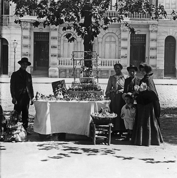 A black and white photo captures an early twentieth century outdoor market scene. A woman and children of different ages gather by a table holding a large amount of figurines of animals and people. 