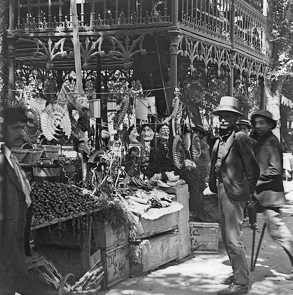 In a black and white photo, a suited man looks into the camera as an outdoor market bustles about him. Masks, fans, and fruits are all arrayed for sale in the stalls.