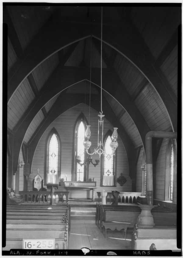 A black and white photo records the pew-lined interior of a small church. Large wooden ribbing lines the ceiling and three windows puncture the apse behind the simple altar.