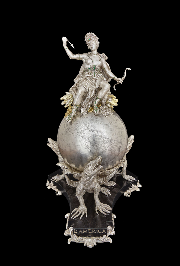 A silver sculpture depicts a bare-chested female allegorical figure sitting atop a globe engraved with a map of the Americas. She holds up an arrow and wears a gem-studded belt, headpiece, and sandals. The globe is held aloft on the backs of alligators.
