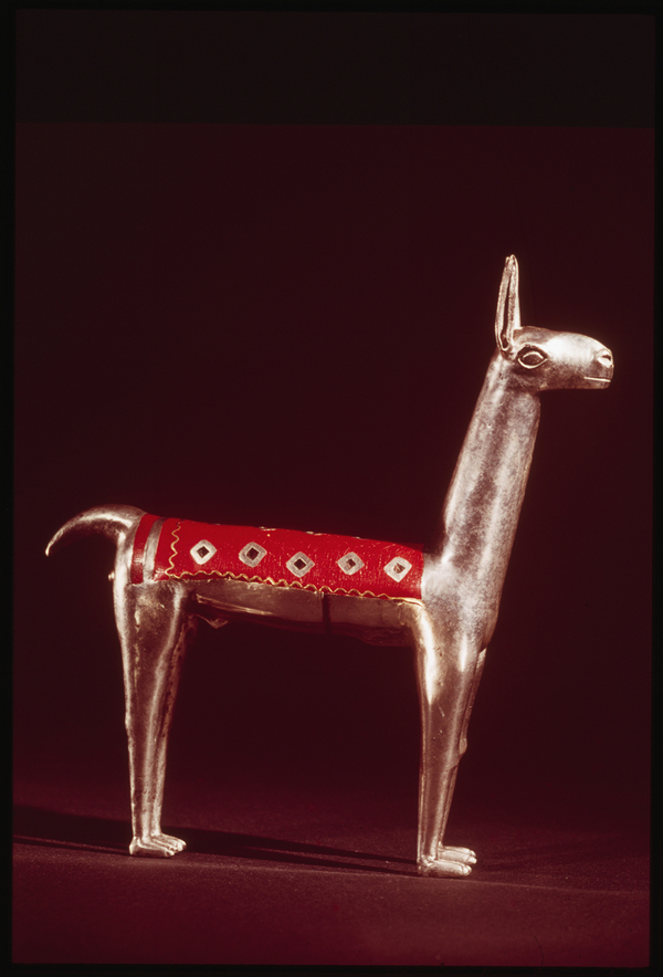 An angular cameloid metal figurine has perky ears and tail. It is dressed with a patterned weaving over its midsection. The covering is red with a golden, wavy border and silver diamond pattern.