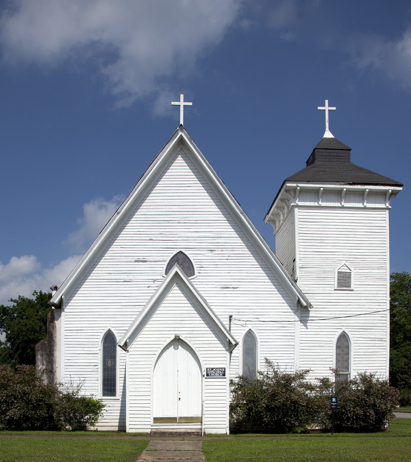 A white church with a steep gable roof and pointed arch windows has a bell tower on the right. A enclosed church porch stands at the entrance. 