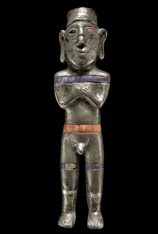 A silver figure with long earlobes and an erect penis clasps his hands to his chest. There is a belt of orange stones set into the figure's waist and one stone in an eye socket. Purple stones belt the chest and encircle each thigh.