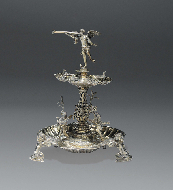 An elaborate bilevel silver centerpiece is topped with a winged angel blowing a horn. The upper dish includes silver birds and the lower dish has frolicking putti. The entire work stands on nude busts that serve as its short legs.