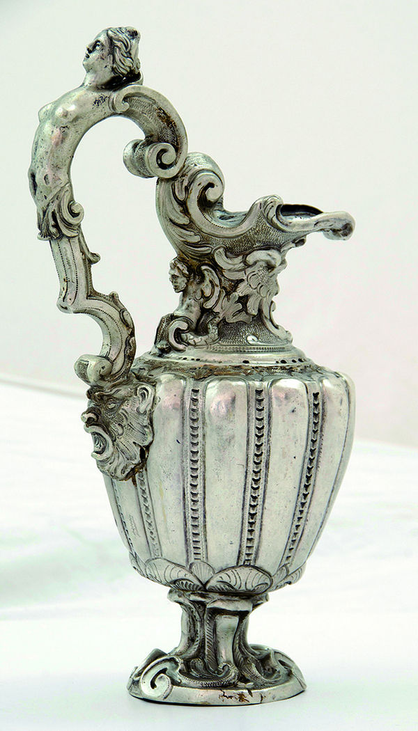 A silver jug is chisel-finished and engraved. Its double volute handle consists of the arched body of a naked woman. A double-tailed male figure leans against the jug's foliage-decorated neck. 