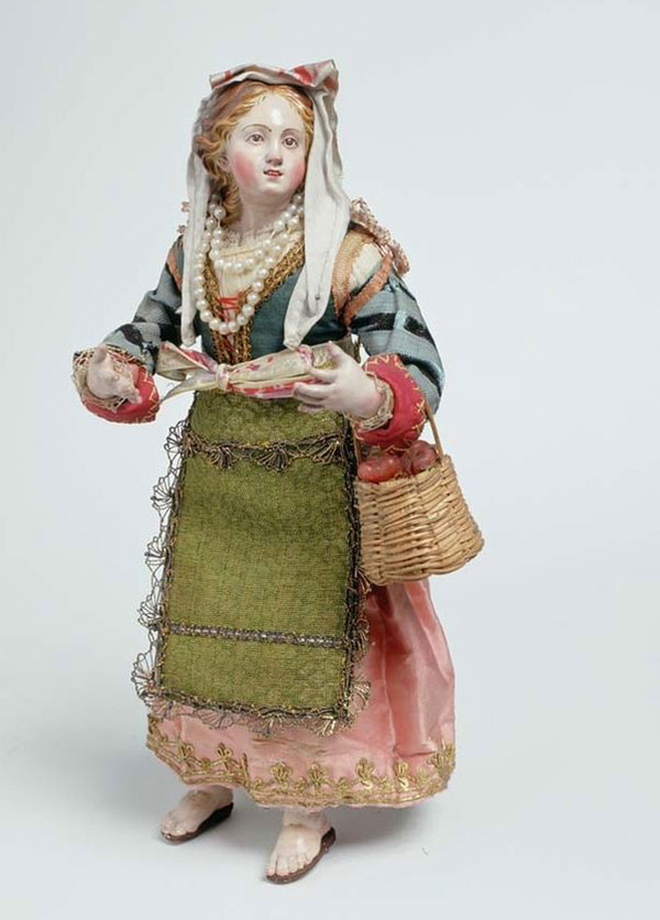 A clay or wood figure depicts a female peasant with pale white skin and rosy cheeks. She has been dressed in a colorful gown with a bow cinched about her waist, pearls, and a veil. She holds a basket of apples on one arm.