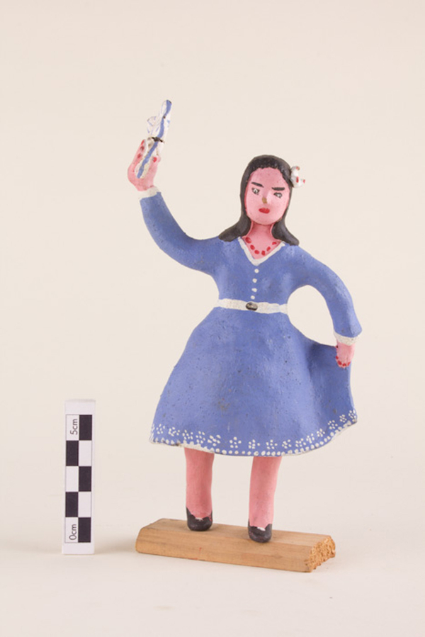 A painted clay figurine depicts a woman dancing in a periwinkle dress. She holds a corner of her dress up in one hand and waves a blue and white object in the air with the other. Her nails have been painted red and match her red necklace.
