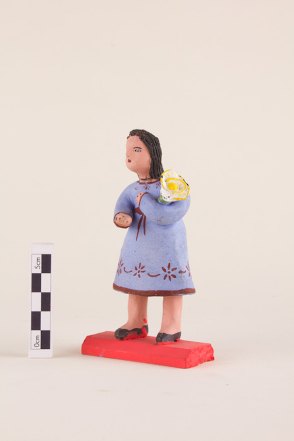 A painted clay figurine depicts a light-skinned woman in a purple dress holding an oversized white and yellow flower in the crook of her arm. She stands on a red platform and her hair has been rendered as thick dark locks.