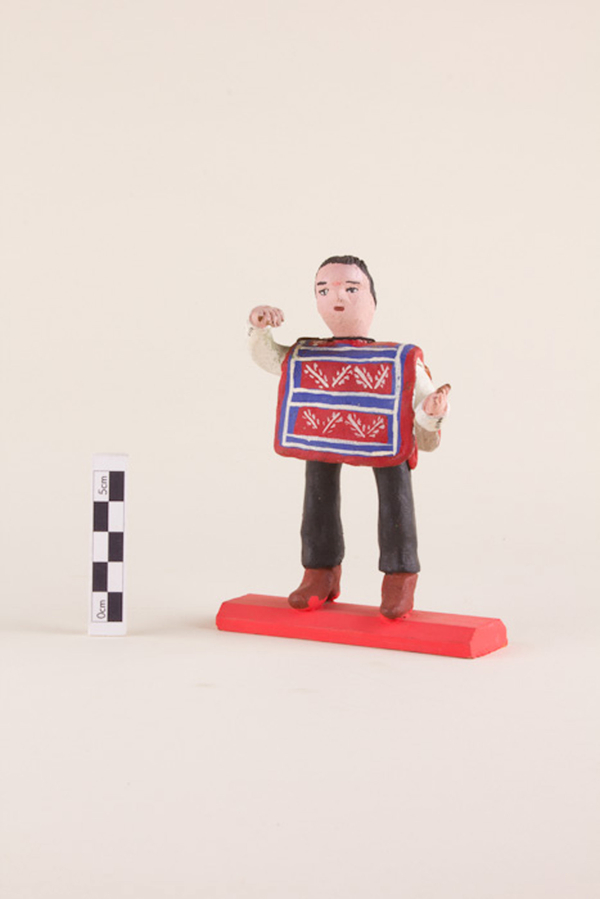 A painted clay figurine depicts a light-skinned huaso wearing a red and blue striped manta with white flower details. He has short brown hair and gestures with his hands. The figure has brown boots and stands on a red platform.