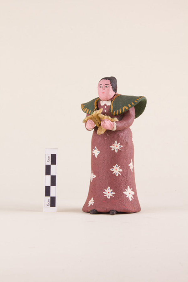 A painted clay figurine depicts a light-skinned woman in a red dress with a white flower pattern. She wears a green shawl that flares out at the shoulders. The woman has light skin and small facial features, and her hair is pulled back into a bun.