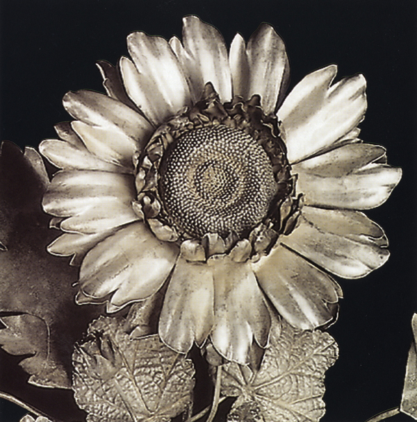 A flower made of silver has two rows of curling petals around a seedy center. Veined silver leaves line the stem.