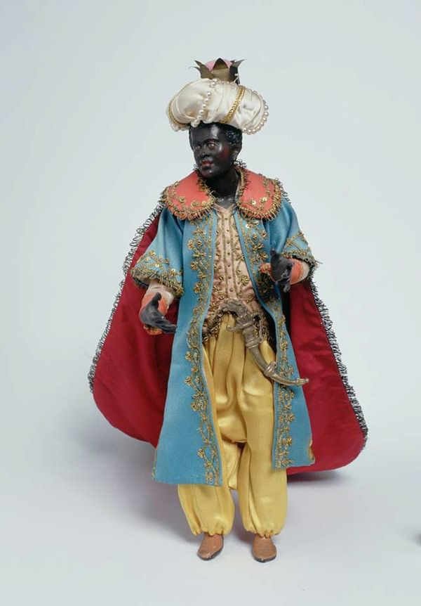 A clay or wood figure of Balthazar has dark skin, curly black hair, and red lips. He has been dressed in a large turban with a crown, a red cape, and rich embroidered robes. A knife hangs at his waist.