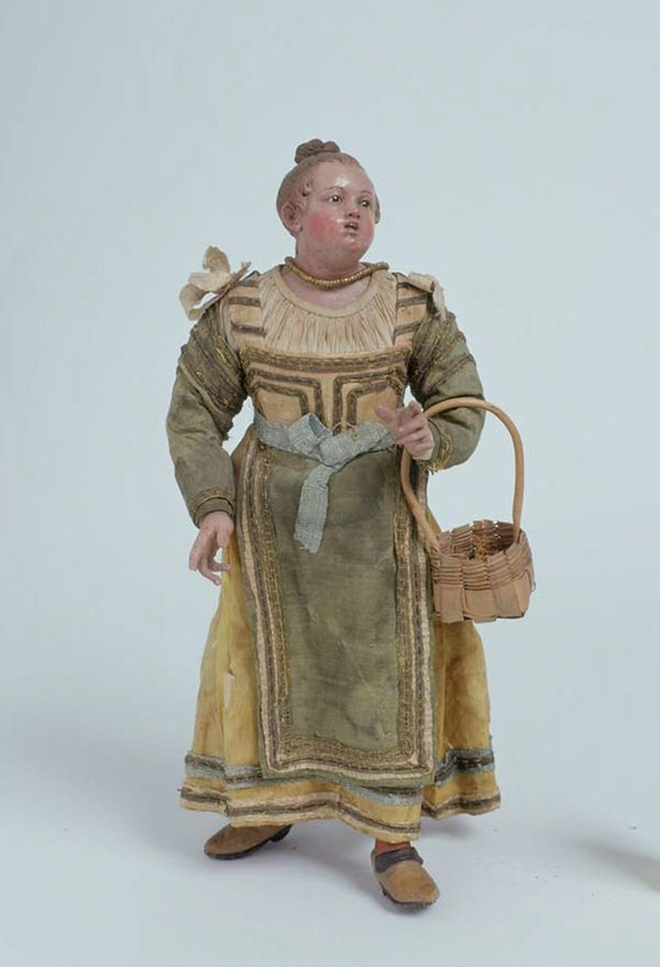 A clay or wood figure depicts a light-skinned female peasant with a surprised expression. She is dressed in a green and yellow dress and wears a gold necklace. A wooden basket is slung over her arm.