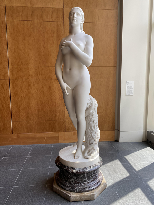 A white marble sculpture of Eve depicts her as an idealized woman standing tall with her arms moving to cover her nakedness. She has a quiet, sad expression with her eyes looking slightly upward.