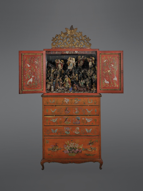 The doors of a red cabinet are open to display an elaborate creche with multiple painted figures. Angels with golden wings fly above Mary, Joseph, and the Christ child. Figures of shepherds, animals, and peasants gather around them.