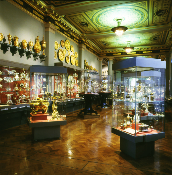Glass cases of silver are on display in an ornate museum gallery. The room has gold moldings and a painted ceiling. More silver works line its shelves and walls.