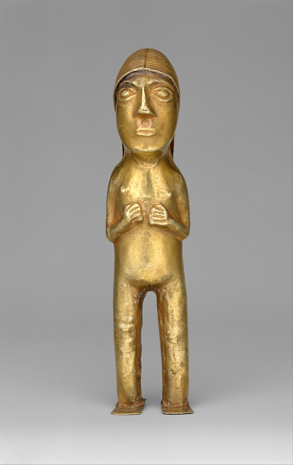 A standing gold figurine with an oversized head, large almond eyes, pursed lips, and slightly raised nipples holds her arms and hands against her chest. 