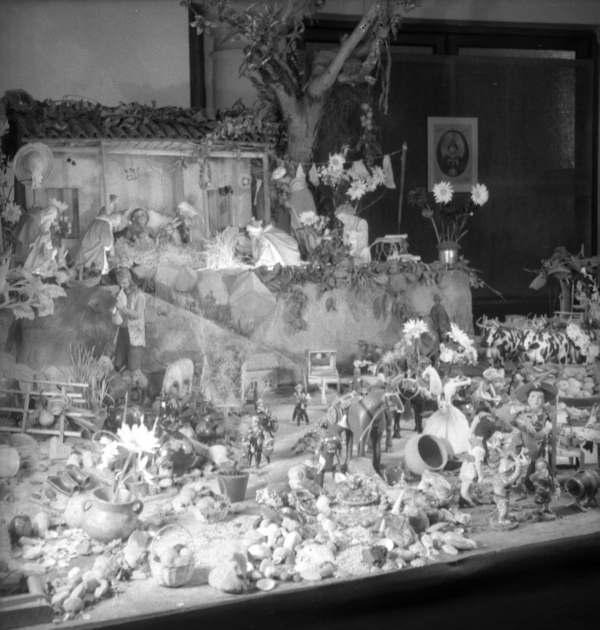 A black and white photo captures some details of an assembled nativity scene. Pots, animals, baskets of food, flowers, and various figurines have been assembled on one level with Christ, Mary, and Joseph have been placed in a stable on a little hill.