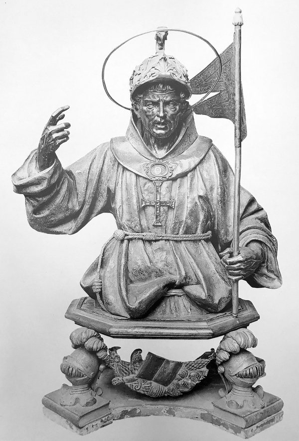 In a silver reliquary bust, San Giovanni da Capistrano is depicted with a stern expression and raised hand as if orating. He holds a pennant in the other hand. The saint is dressed in Franciscan robes and has a helmet-like cap and halo on his head.