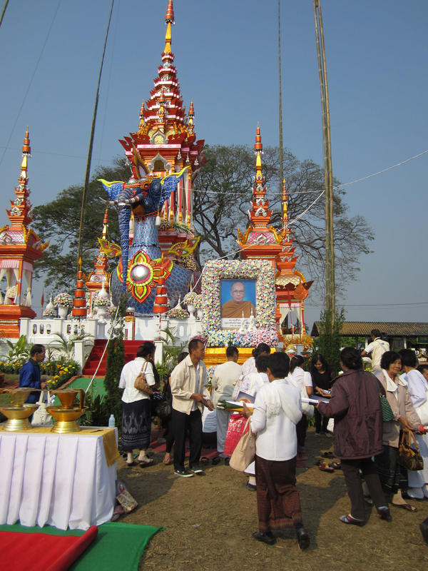 Thai people of different ages and genders gather around a brightly colored prasat nok hatsadiling structure. It has a blue elephant head atop a bird. An image of a monk framed by flowers stands next to the structure.