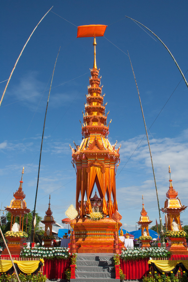 A golden prasat sop with open-sides is built atop a decorative plinth for the body. It has bright hanging curtains and a layered roof on top that culminates in a gold spire. The roof eaves are adorned with flames.