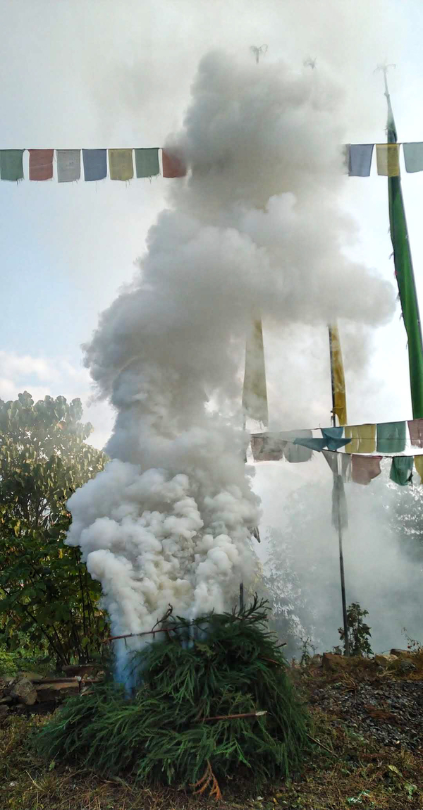 Smoke billows out from a pile of lit tree branches constructed outside. Colorful prayer flags are strung up in the background.