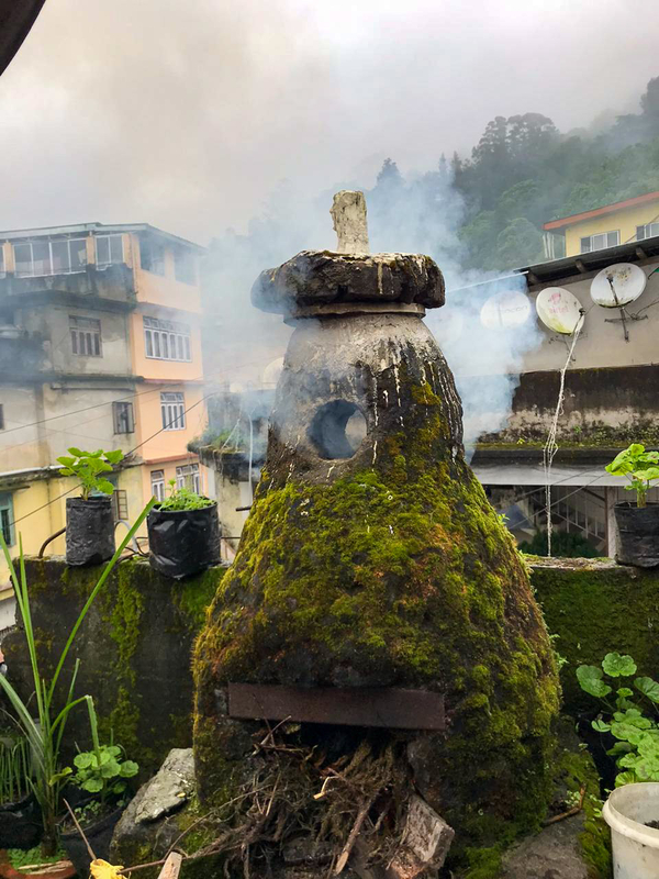 A mound-like repository has a section at the bottom filled with lit twigs and a circular opening at the top for smoke's escape. Moss covers the vessel and it sits on the top of a building.