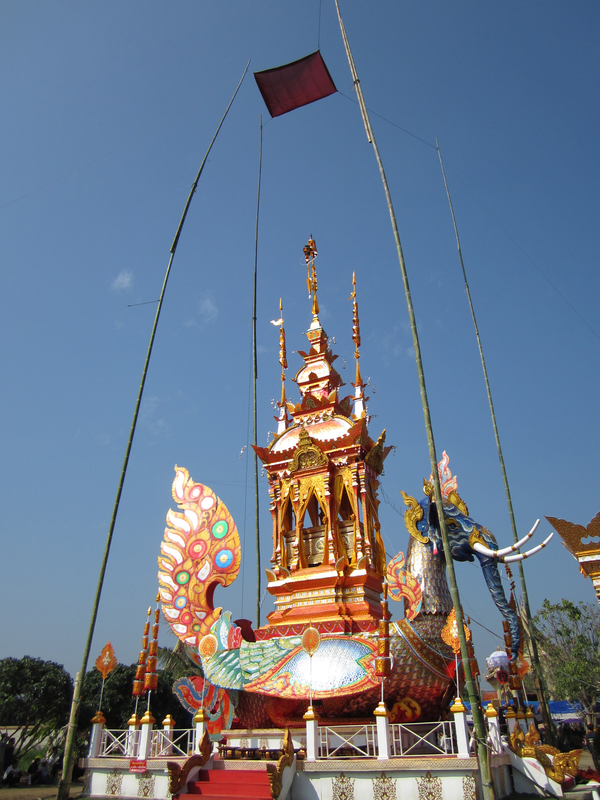 In an elaborate cremation construction, a gold, open-sided structure with a round roof and towering spires sits atop a massive and colorful, elephant-headed bird. 