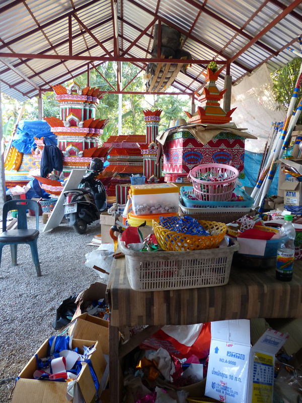 Baskets of colored papers and other craft items are arrayed on tables under a pavilion. A bright red prasat sop structure in the process of being assembled is visible in the background. 
