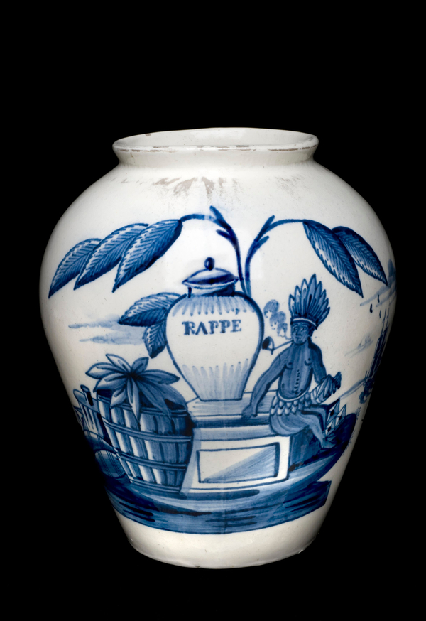 A white ceramic snuff jar is decorated with a blue scene of a native man smoking a pipe and sitting on the platform of a large jar inscribed "RAPPE."  Barrels of tobacco are shown nearby while a leafy plant looms over the scene.